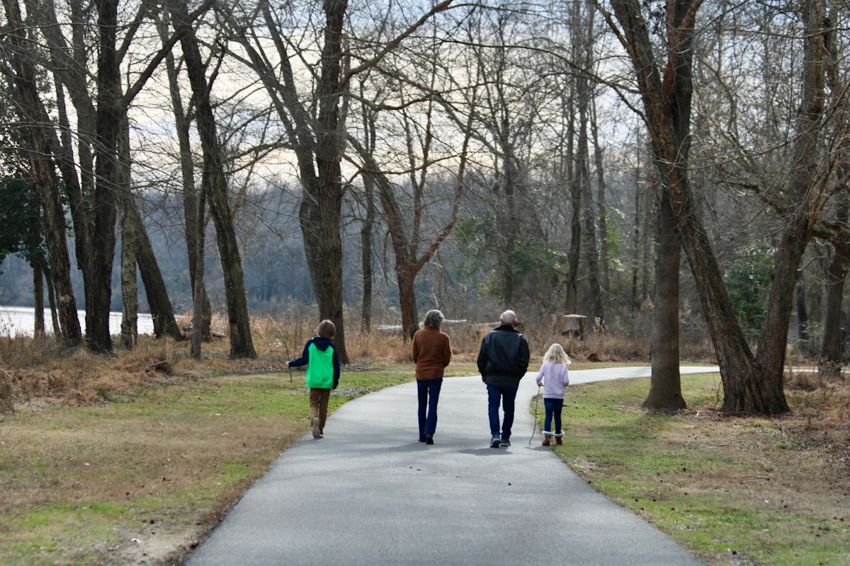 Family of four seen from behind walking a curved paved path through leafless trees beside a river on an overcast day